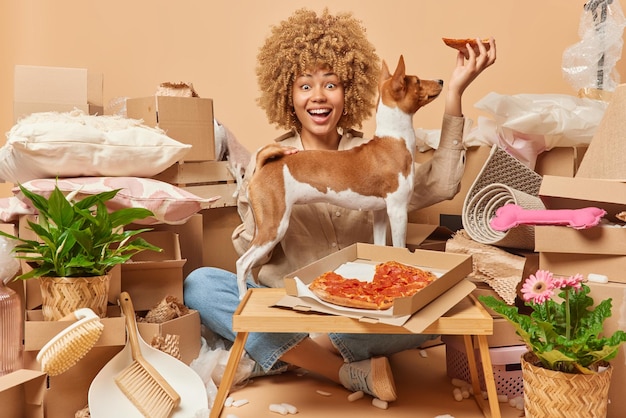 Happy curly haired european woman eats pizza together with pet\
moves in new apartment relocates surrounded by boxes full of\
belongings looks gladfully at camera new home and moving\
concept