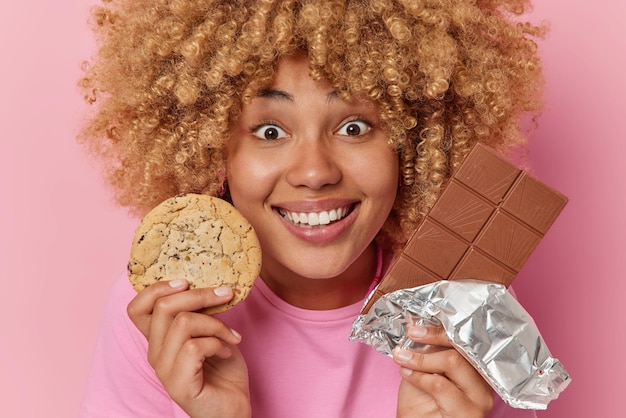 happy curly haired european woman holds chocolate bar and cookie enjoys sweet life smiles happily prefers eating unhealthy sugary food dressed in casual t shirt isolated over pink background