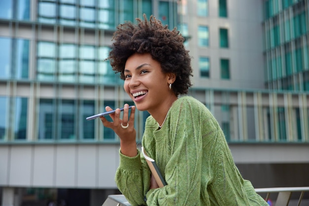 Happy curly haired woman dressed in green jumper holds notepad uses voice command recorder on smartphone translates words with application stands against blurred city background records message