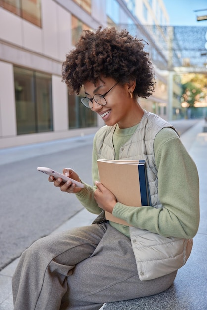 Happy curly haired woman uses mobile application for browsing media content reads receieved message holds textbooks wears jumper vest and trousers sits outdoors during sunny day Technology concept