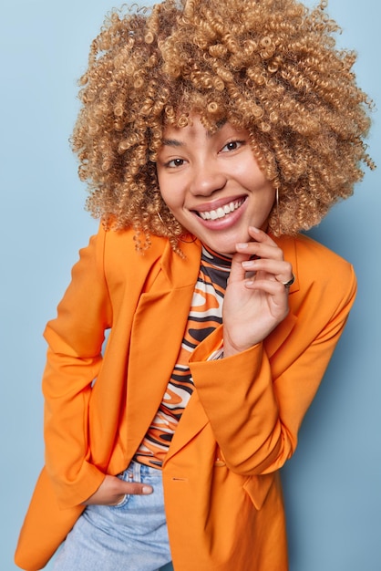 Happy curly haired young european woman smiles broadly dressed\
in fashionable orange jacket and jeans keeps hand in pocket keeps\
hand on face being in good mood isolated over blue background