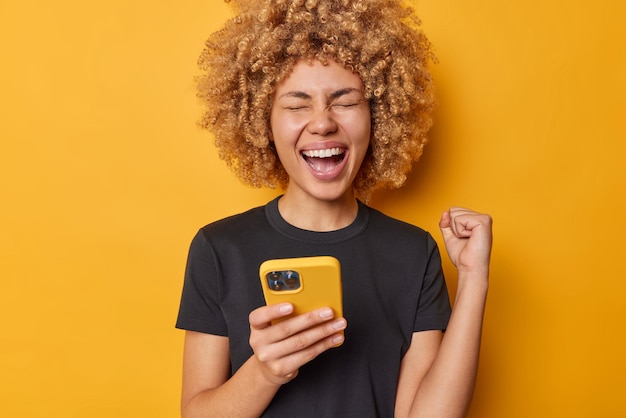 Foto felice giovane donna dai capelli ricci stringe il pugno gioisce del successo tiene lo smartphone moderno riceve un messaggio sulla ricompensa indossa una maglietta nera casual isolata su sfondo giallo fa il gesto del vincitore