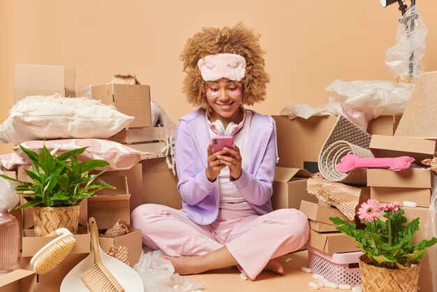 Happy curly haired young woman finds new apartment for renting\
surfs internet via smartphones undergoes beauty procedures packs\
belongings in carton boxes makes orders online ownership\
concept