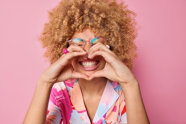 Happy curly haired young woman makes heart gesture over mouth smiles toothily wears transparent spectacles and colorful shirt says i love you isolated over pink background body language concept