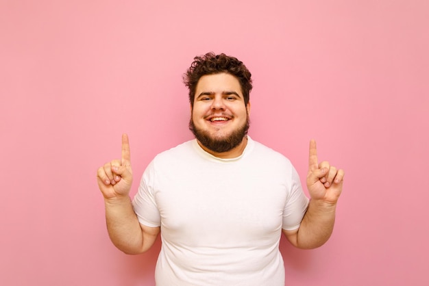 ピンクの背景に白いTシャツの立っている幸せな巻き毛の太りすぎの男はカメラに見えます