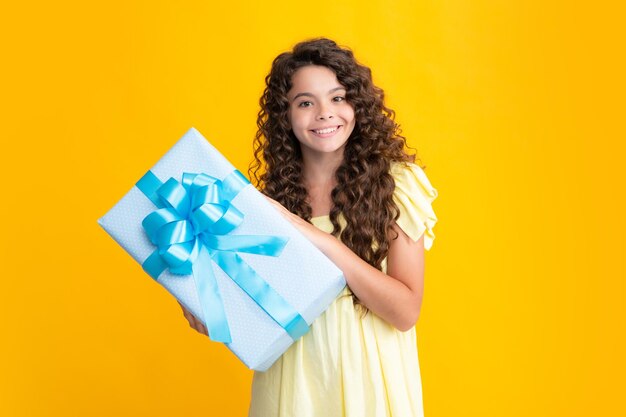 Happy curly teenager portrait Teenager child holding gift box on yellow isolated background Gift for kids birthday Smiling girl