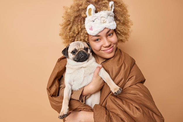 Happy curly woman goingt to take nap with pet wrapped in blanket embraces pug dog and smiles at camera enjoys comfort and domestic atmosphere during rainy day isolated over brown background.