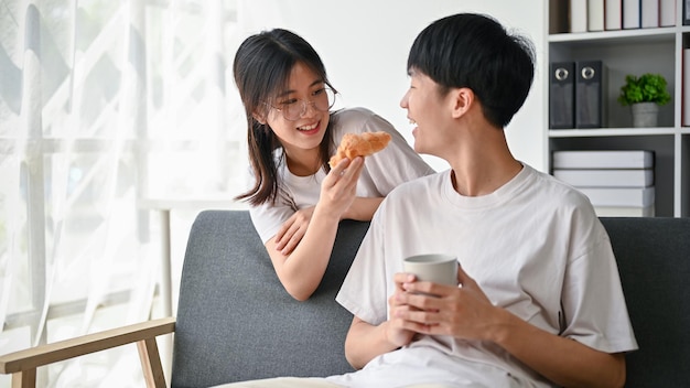 행복하고 귀여운 아시아 여자 친구가 남자 친구에게 맛있는 크루아상을 주고 놀고 있습니다.