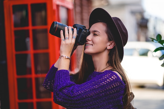 Fotografo attraente felice sveglio della giovane donna in cappello con la macchina fotografica digitale della foto del dslr durante la presa delle foto nella città nel tempo di viaggio