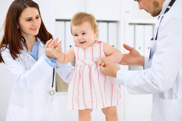 Happy cute baby at health exam at doctor's office Toddler girl is standing while have been keeping by two doctors