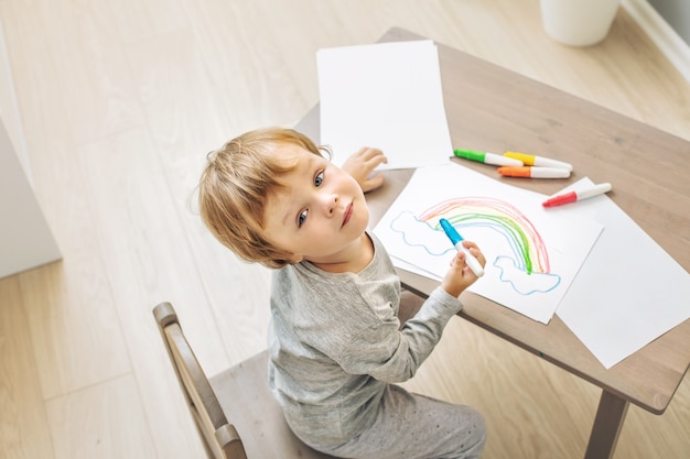 Foto bambino carino e bello felice che sorride a casa disegnando al tavolo nella scuola materna