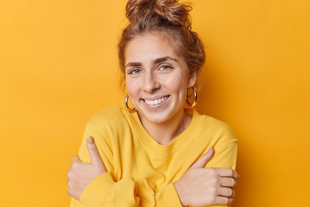 Happy cute blue eyed european woman with combed hair smiles toothily wears soft jumper and earrings feels comfortable hugs herself isolated over vivid yellow background self emotions concept