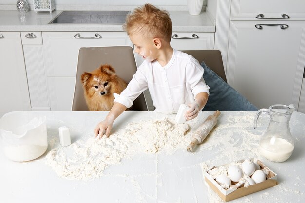Happy cute boy covered in flour cooks with a red dog in the kitchen