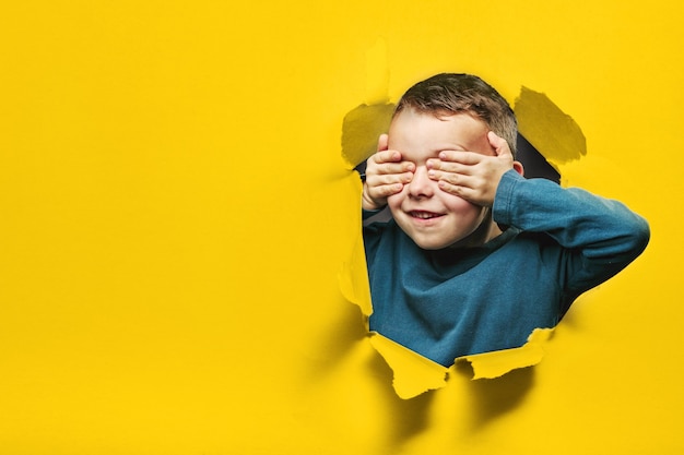 Photo happy cute boy is having fun played on black background wall. climbs through a hole in the paper. bright photo of a boy.