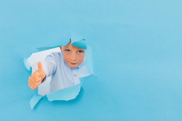 Happy cute boy is having fun played on blue background wall climbs through a hole in the paper