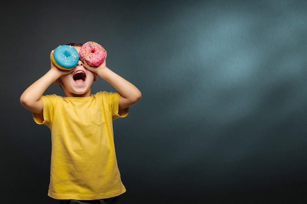 Happy cute boy is having fun played with donuts on black background wall.