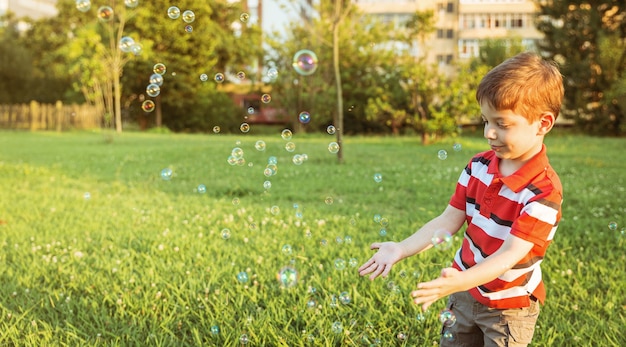 Счастливый милый мальчик играет, чтобы поймать мыльные пузыри в парке