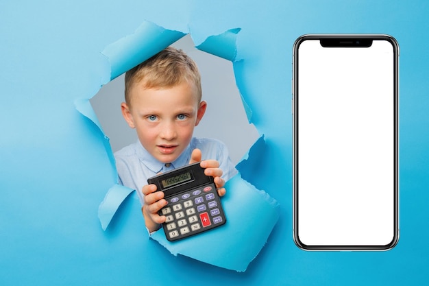 Happy cute businessboy is having fun and holding calculator in hand on blue background wall climbs through a hole in the paper near huge mobile phone