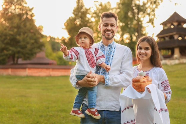 Happy cute family in embroidered Ukrainian shirts with korovai bread on sunny day Space for text