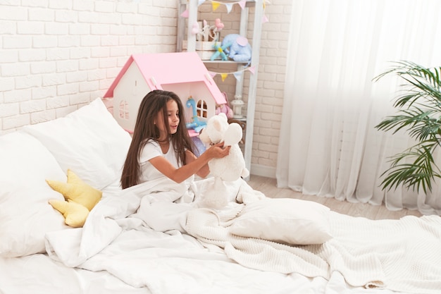 幸せなかわいい女の子が彼女の子供部屋のベッドに座って、テディベアと遊ぶ