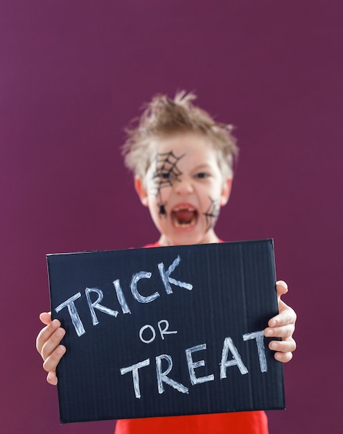 写真 トリックオアトリートというテキストの黒板を持って、ハロウィーンの化粧をした幸せなかわいい子供。ハロウィーンのコンセプト。