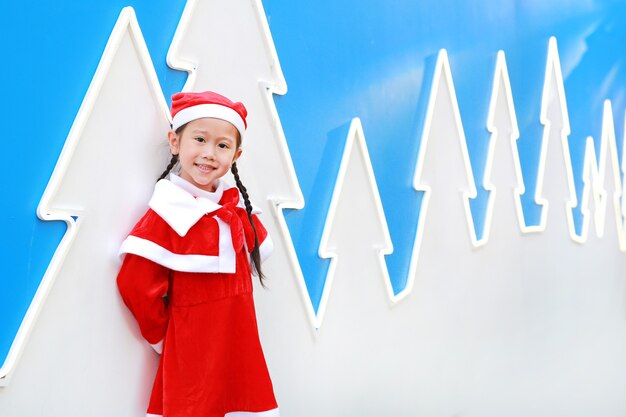 Фото Счастливая милая маленькая азиатская девушка ребенка в костюме санты около рождественской елки и предпосылки