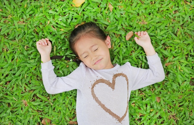 Happy cute little Asian child girl lying on green lawn. Smiling and closed her eyes.