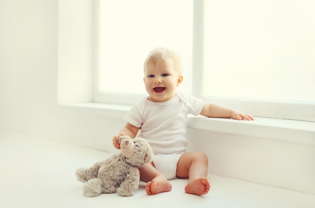 Happy cute little baby playing with teddy bear toy in white room at home