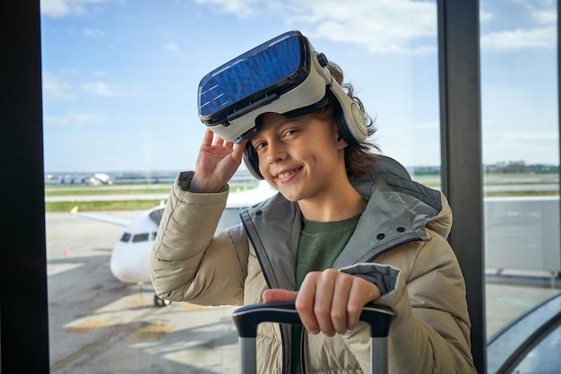 Happy cute little boy in warm jacket taking off VR headset and smiling while standing near window in airport with suitcase and looking at camera before flight