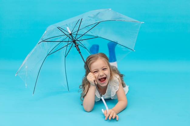 A happy cute little girl in blue rubber boots and a cotton white dress holding an umbrella lying on a blue background in the studio laughing smiling and fooling around a place for text