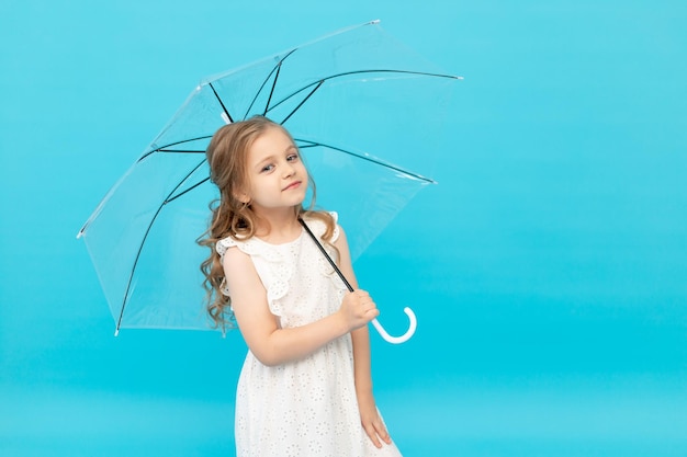 Happy cute little girl in a cotton white dress holding a transparent umbrella on a blue background in the studio and smiling and fooling around space for text