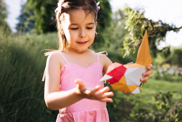 Happy cute little girl holding paper colorful bird in the park Child playing with a bird toy outdoors Ecology and environment concept Childhood Kid learning about caring and protecting the nature