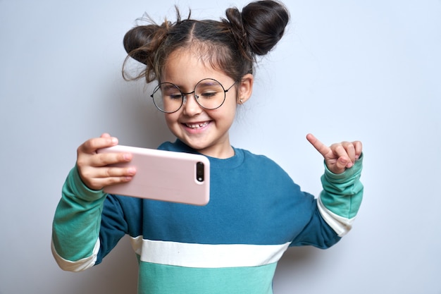 Happy cute little girl isolated on white studio background with mobile phone in hands, child holding smartphone smiling laughing