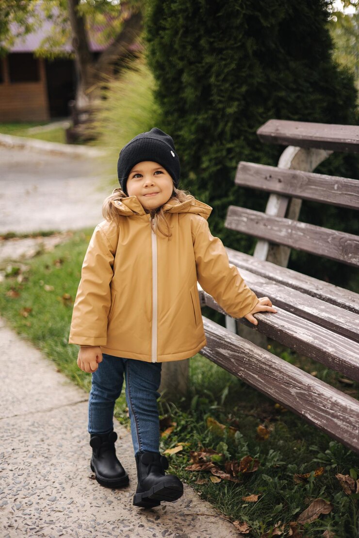 Photo happy cute little girl in jacket and hat posing to photographer outdoor by the bench little model
