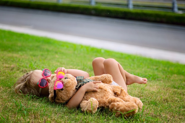 La bambina sveglia felice che si trova sull'erba con il suo orsacchiotto gioca in occhiali da sole