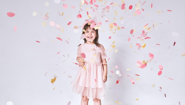 Happy cute little girl wearing pink dress in tulle with princess crown on head isolated on white background playing with confetti Joyful pretty little girl celebrating her birthday party having fun