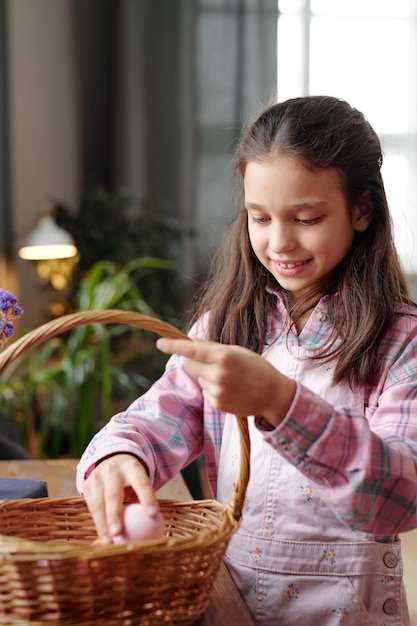 Happy cute little girl with toothy smile standing by table while putting painted Easter egg into basket during preparation for holiday