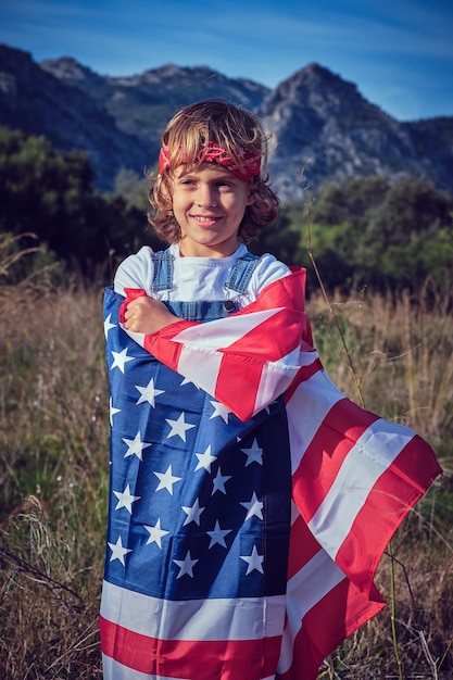 Happy cute preteen boy standing in grassy meadow wrapped in national flag of United States of America