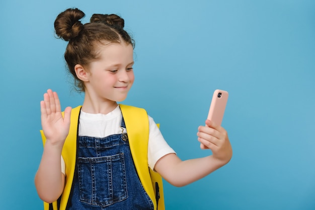 Happy cute schoolgirl with backpack holding phone, waving hand, video calling friend, family or school teacher during virtual meeting remote distance learning posing isolated on blue studio background