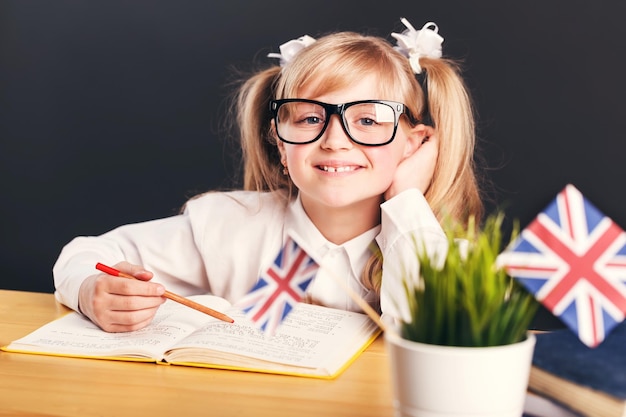 행복한 귀여운 웃는 소녀는 어두운 배경 앞에서 책으로 영어를 배우는 똑똑한 안경을 쓰고 있다