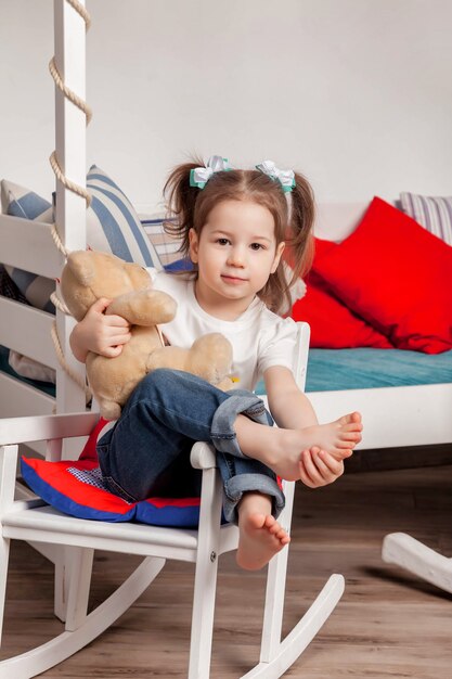 Happy cute three-year-old girl sitting in her children's room and hugs Teddy bear. Lovely lucky little child with Teddy bear on chair in home and looking at camera. Atmospheric baby's family moments