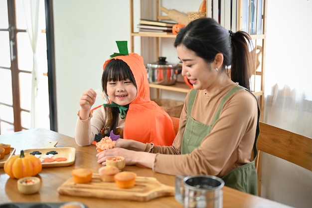 幸せでかわいい若いアジアの女の子は、お母さんがハロウィーンのカップケーキを飾るのを楽しんでいます