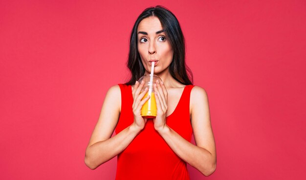 Happy cute young smiling woman with long curly hair in red swimsuit posing over pink background with orange fresh or juice in hand Summer drink