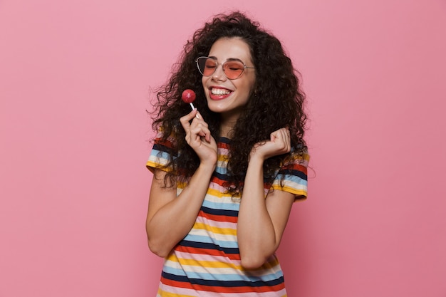Happy cute young woman posing isolated on pink eat candy lollipop.