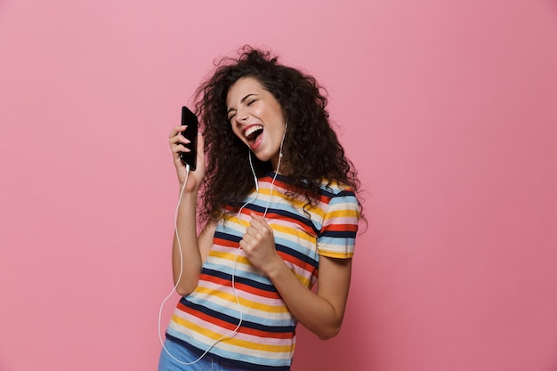 Felice carino giovane donna in posa isolata sul rosa utilizzando la musica d'ascolto del telefono cellulare.