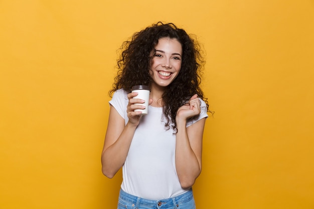 Happy cute young woman posing isolated on yellow holding cup of coffee.