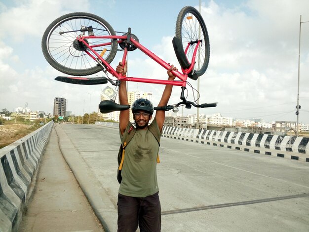 Photo happy cyclist carrying bicycle while standing on highway in city against sky