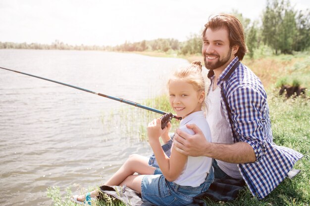 Photo happy dad and daughter are sitting on grass near water and looking at camera. he is huging her and holding fish-rod in right hand. they are fishing.