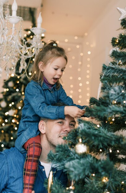 행복한 아빠와 딸이 크리스마스 트리를 장식합니다 어린 소녀가 아빠의 어깨에 앉아 있습니다