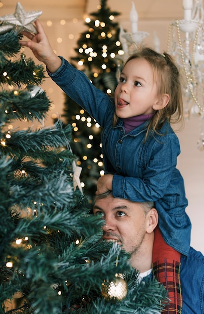 幸せなお父さんと娘がクリスマスツリーを飾ります小さな女の子がパパの肩に座っています